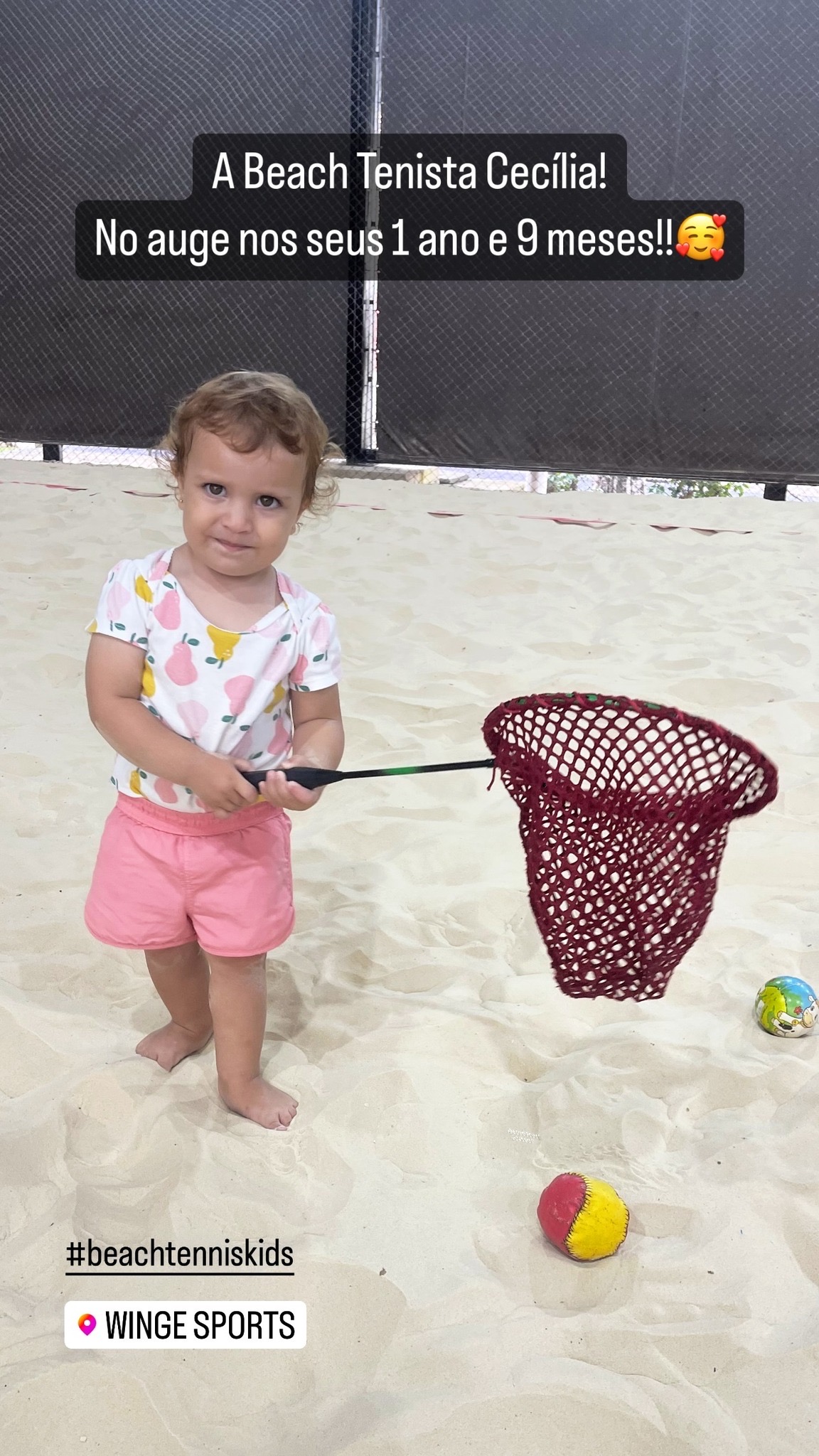 Qual a idade ideal para começar no Beach Tennis Infantil? 1 Bebê Cecília, de 1 ano e 9 meses, brincando com raquete e rede de beach tennis na areia, demonstrando os primeiros contatos infantis com o esporte.