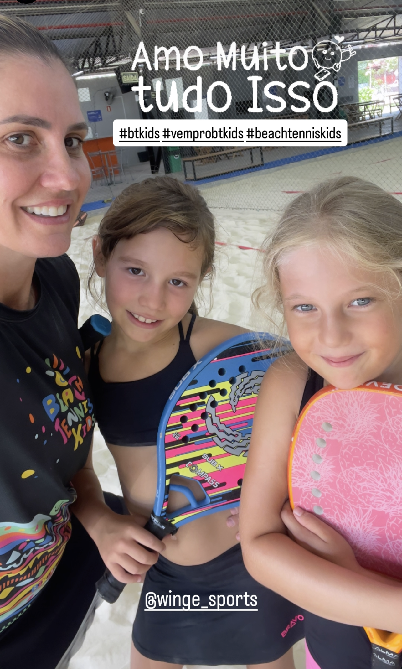 10 Benefícios Essenciais do Beach Tennis Kids: Saúde e Emoção em Quadra 3 Close-up de professora e duas alunas sorrindo, segurando raquetes de Beach Tennis Kids, em um ambiente de quadra de areia. Foco na amizade e na interação positiva