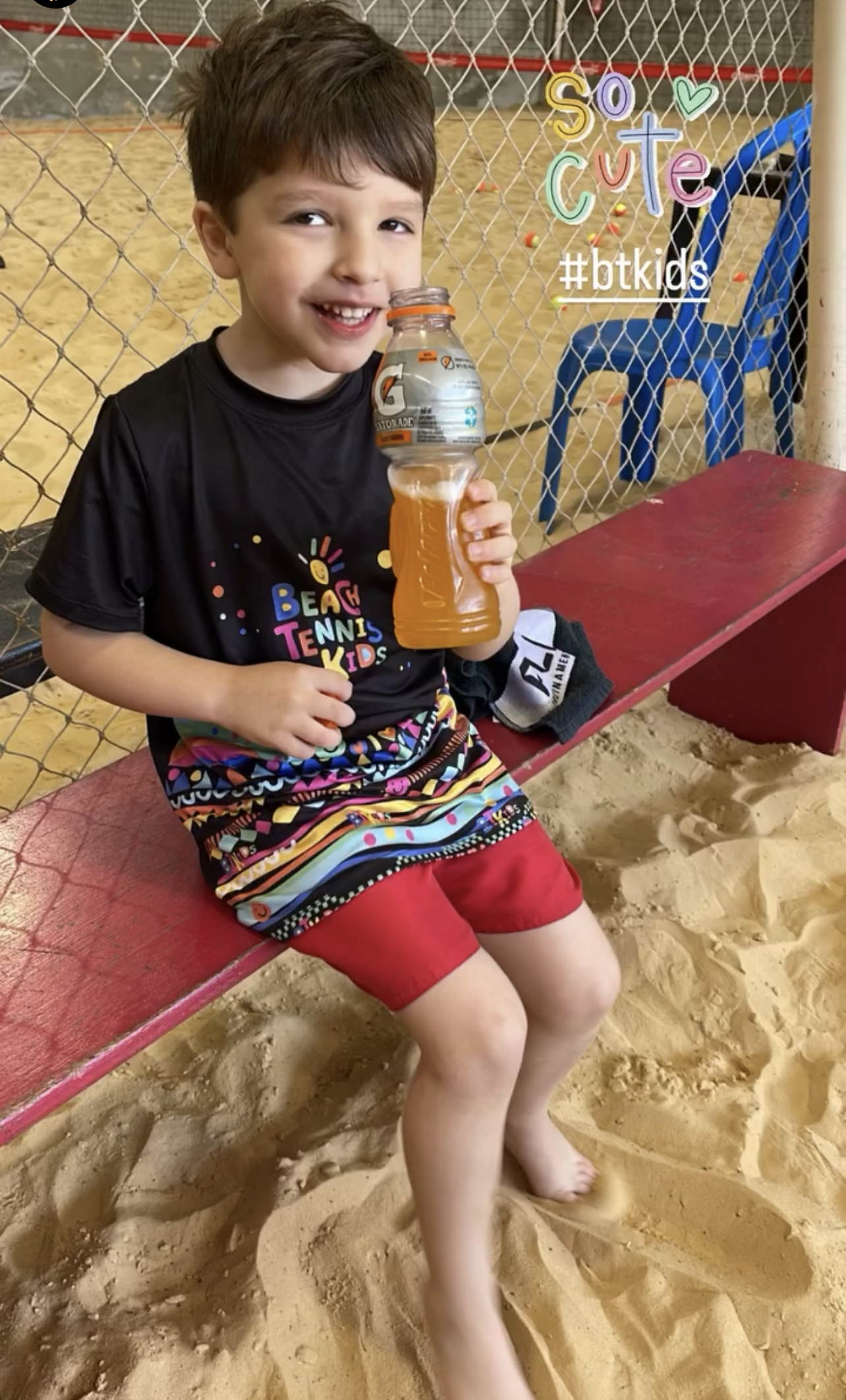 10 Benefícios Essenciais do Beach Tennis Kids: Saúde e Emoção em Quadra 1 Criança sorrindo, sentada na areia de uma quadra de Beach Tennis Kids, segurando uma garrafa de isotônico, após o treino, demonstrando hidratação e alegria pós-atividade física.